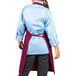 A woman wearing a blue shirt and a burgundy Uncommon Chef bib apron with three pockets.