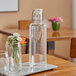 An American Metalcraft clear acrylic hinged water bottle with a glass of water on a table.