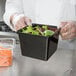 A chef in gloves holding a black square melamine crock filled with salad.