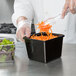 A person pouring carrots into a black square melamine crock.