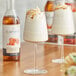 A close up of a Capora Salted Caramel Flavoring Syrup bottle on a table with two glasses of white and brown liquid.