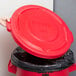 A hand placing a Rubbermaid red lid on a trash can.