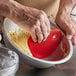 A person's hand using a red silicone bowl scraper to mix dough in a bowl.