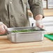 A man in a chef's coat holding a stainless steel Choice 1/2 size steam table pan filled with green beans.