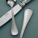 A close-up of a Sant'Andrea Bellini stainless steel round bowl soup spoon and a knife on a table.