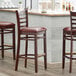 Three Lancaster Table & Seating mahogany wood bar stools with burgundy vinyl seats and ladder backs at a bar counter.