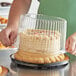 A person holding a cake with a clear plastic dome lid.