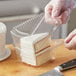 A hand in a plastic glove cutting a piece of cake in a clear plastic container.