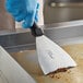 A gloved hand with a blue glove and a black-handled Ateco stainless steel chocolate scraper using a spatula over a metal pan.