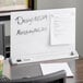 A white Quartet frameless magnetic glass dry erase board on a desk with a piece of paper pinned to it.