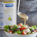 A spoon pouring Hellmann's Greek Vinaigrette dressing onto a bowl of salad.