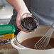 A person using a whisk to stir brown liquid in a bowl.