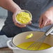 A person stirring yellow liquid in a white pot with a spoon.