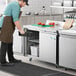 An aproned man standing next to an Avantco stainless steel worktop freezer.