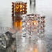 A clear glass Hollowick faceted cube votive with a lit candle inside on a table.