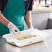 A person in a green apron using gloves to put a lemon in a white Vigor food storage container.