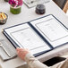 A hand holding a blue vinyl menu cover with gold corners on a white tablecloth.