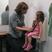 A woman in a grey shirt helping a child in a pink shirt sit in a Koala Kare child protection seat.