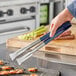 A person using Vollrath VersaGrip tongs with blue handles to grill vegetables.