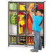 A young boy in a blue shirt standing in front of a Rainbow Accents black and gray coat locker holding a green bag.