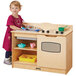 A girl in a pink dress standing next to a Jonti-Craft toddler kitchen with a sink and stove.