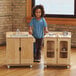 A little girl standing next to a Jonti-Craft TrueModern wooden play kitchen with a pan and pot on top.