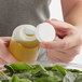 A hand holding a Tablecraft white plastic bottle cap opening a bottle of liquid over a bowl of spinach.