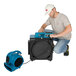 A man kneeling on the floor next to a blue XPOWER air scrubber with a power outlet.