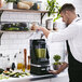 A man in a kitchen pouring green liquid into a KitchenAid commercial blender on a counter.