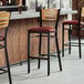 A row of Lancaster Table & Seating bar stools with burgundy vinyl seats and natural wood backs at a bar counter.