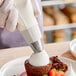 A person using an Ateco plain piping tip to pipe cream on a brown cake.