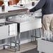 A man in a blue shirt and khaki pants reaching for glasses on a Regency stainless steel underbar drainboard.