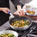 A person cooking food in a Choice aluminum non-stick fry pan with a blue silicone handle.