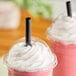 A black pointed smoothie straw inserted into a pink smoothie with whipped cream in a clear plastic cup.