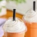 A clear plastic cup with a dome lid and a black smoothie straw, filled with an orange beverage topped with whipped cream.