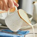 A person pouring cream from an Acopa Keystone Vanilla Bean stoneware creamer into a white cup.