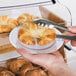 A person holding a white plastic plate with silver bands full of croissants.