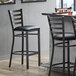 A Lancaster Table & Seating distressed copper ladder back bar stool with a black vinyl padded seat next to a table.