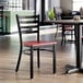 A black ladder back chair with a mahogany wood seat and a distressed copper finish, shown in a restaurant setting.