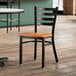 A Lancaster Table & Seating black finish ladder back chair with a cherry wood seat sits in front of a table in a restaurant.