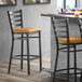 A Lancaster Table & Seating black ladder back bar stool with a light brown cushion next to a table.