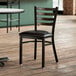 A Lancaster Table & Seating distressed copper ladder back chair with black vinyl seat pad on a wood floor in a restaurant.