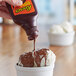A hand pours REESE'S chocolate and peanut butter shell topping onto a bowl of ice cream.