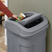 A hand putting a paper cup into a Toter Slimline trash can with a graystone swing lid.