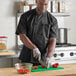 A man wearing a black Uncommon Chef short sleeve chef coat cutting vegetables.