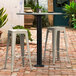 A black Lancaster Table & Seating Excalibur bolt down outdoor table base with a white bar stool on a brick patio.