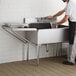 A man in a white shirt and black apron washing a glass in a stainless steel sink with a Regency detachable drainboard.