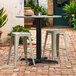 A Lancaster Table & Seating Excalibur black outdoor table base with a plate on it, surrounded by white stools on a brick patio.