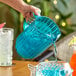 A person pouring a blue drink into a turquoise plastic pitcher shaped like a fish.