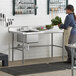 A woman standing in a kitchen with a Regency stainless steel work table with a sink and cross bracing.